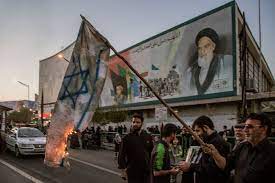 مردان در یک خیابان شهر با یک ماشین پلیس در نزدیکی آن ایستاده و پرچم اسرائیل را می سوزانند. پشت سر آنها یک بیلبورد بزرگ از مردی با ریش سفید و کلاه سیاه دیده می شود.